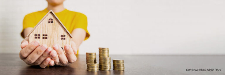 Bausparen - Person mit gelben Pullover hält Haus aus Holz in der Hand, daneben auf einem Tisch liegen Geldmünzen.